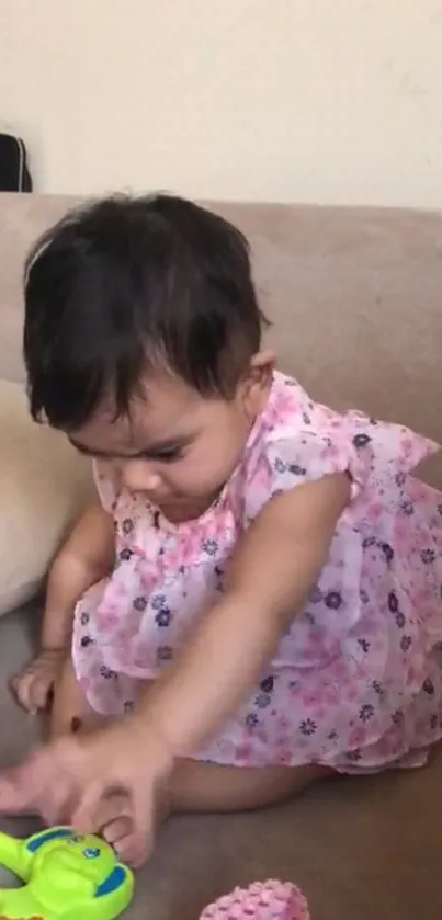 Baby in pink dress plays with toy on beige couch.