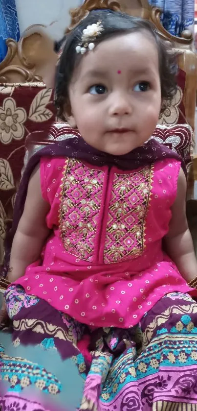 Adorable baby in pink embroidered outfit sitting on a stylish chair.