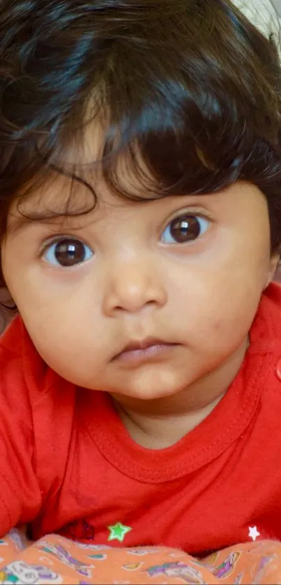 Adorable baby with large eyes in red shirt, looking curious.