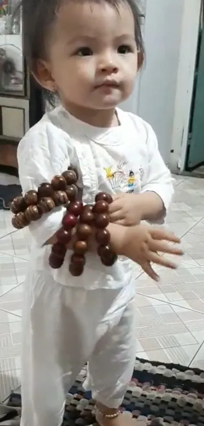 Adorable baby in white holding beads, standing on tiled floor.