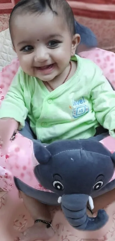 Smiling baby sitting with elephant toy in a pink-themed background.