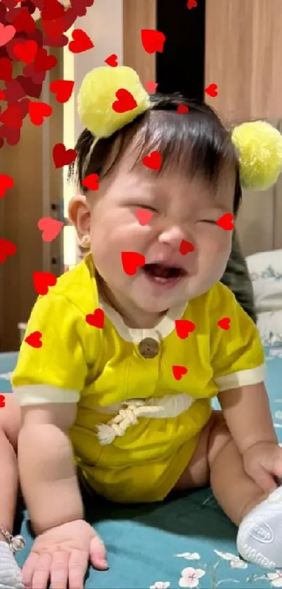 Smiling baby with heart emojis in yellow outfit on bed.