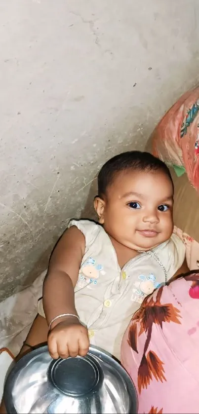 Smiling baby holding a toy bowl next to a colorful pillow.