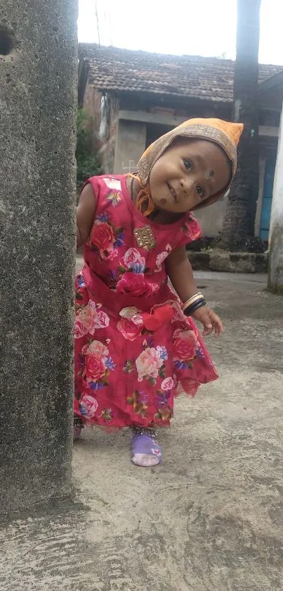 Adorable child in pink floral dress peeking from behind a rustic wall.