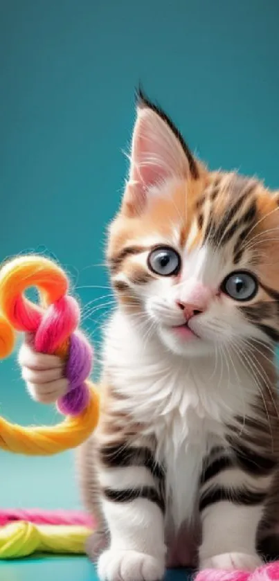 Adorable kitten playing with colorful yarn on a teal background.