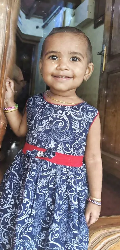 Smiling child in a patterned blue dress by a wooden door.