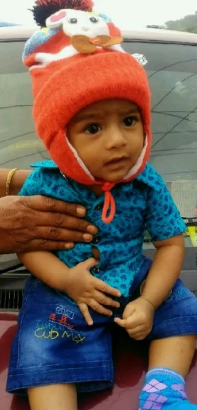 Adorable toddler in bright winter hat sitting on a car.