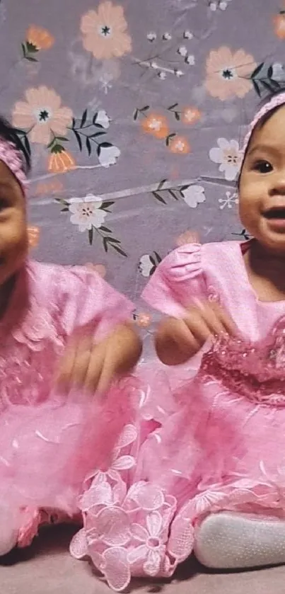 Adorable twin baby girls in pink dresses with floral background.