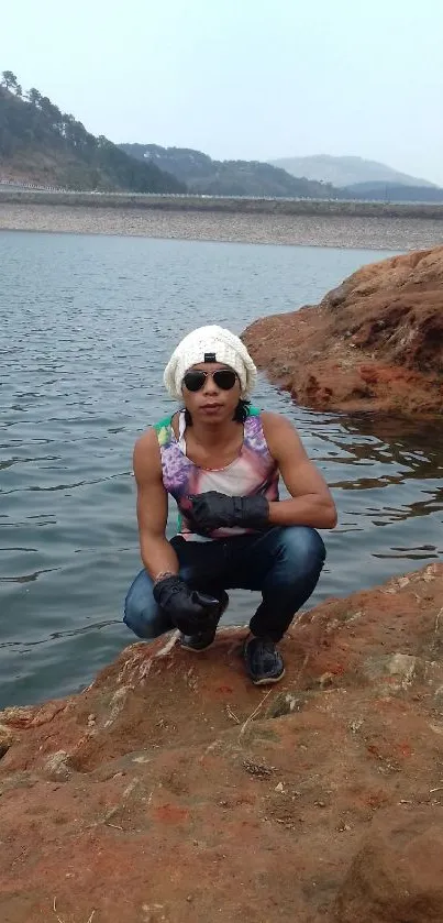 Adventurer crouching by rocky lakeside under cloudy sky.