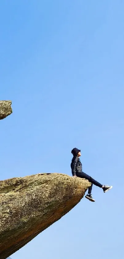 Person sitting on a rocky cliff edge under a clear blue sky.