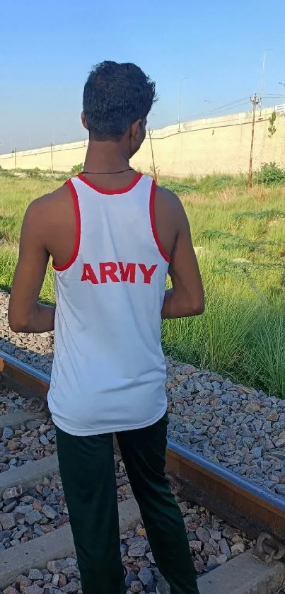 Runner in an Army tank top on railway tracks under a bright blue sky.