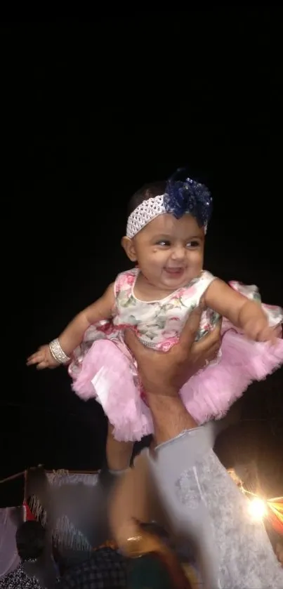 A joyful baby lifted high wearing a floral dress and smiling at an event.