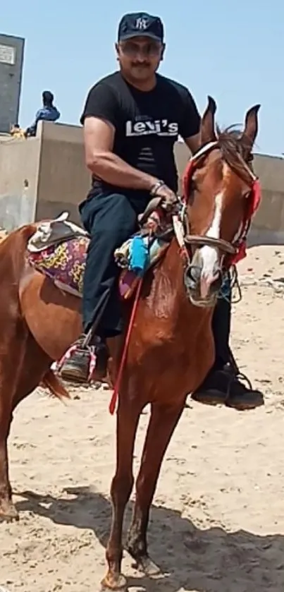 Man riding a brown horse on a sandy beach under a clear blue sky.