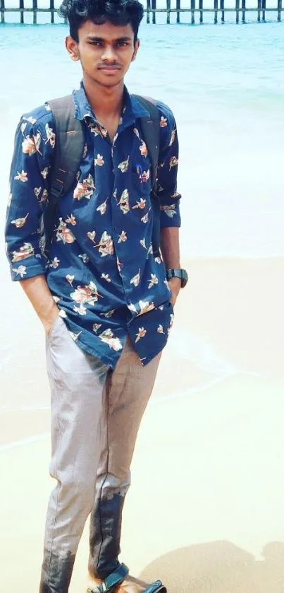 Young man in floral shirt standing on a beach with ocean background.