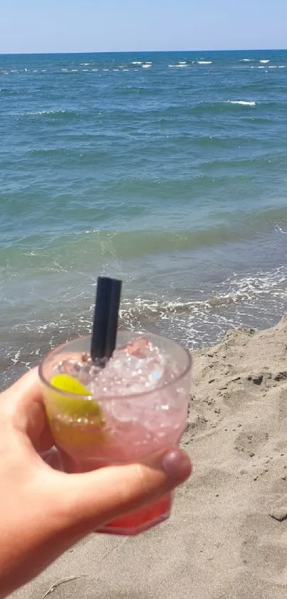 Hand holding a drink by the ocean with sandy beach in view.