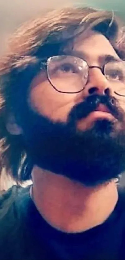 A bearded man with glasses, wearing a black shirt, looks upwards against a blurred background.