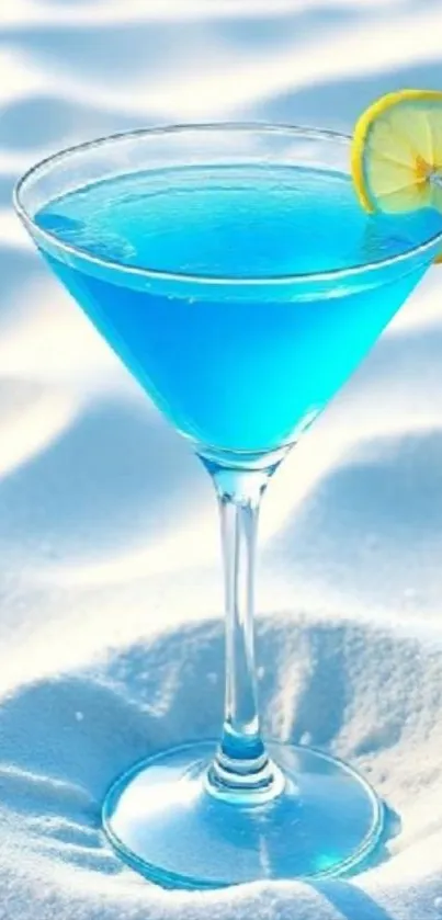 Blue cocktail in a martini glass on white sand beach.