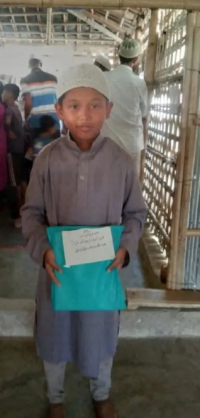 Young boy in rustic setting holding a blue book, surrounded by rural community.