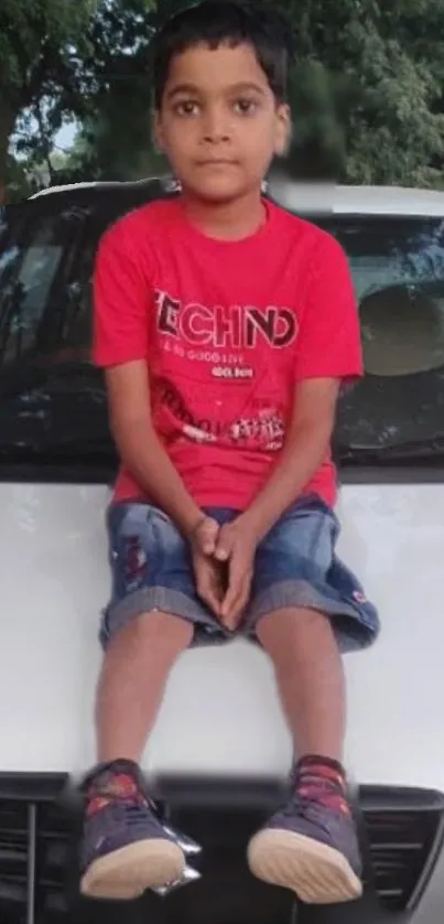 Young boy in red shirt sitting on a white car hood with trees in the background.