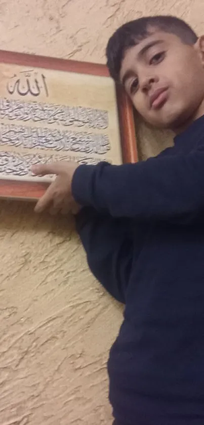 Boy holding framed Islamic calligraphy on textured wall.