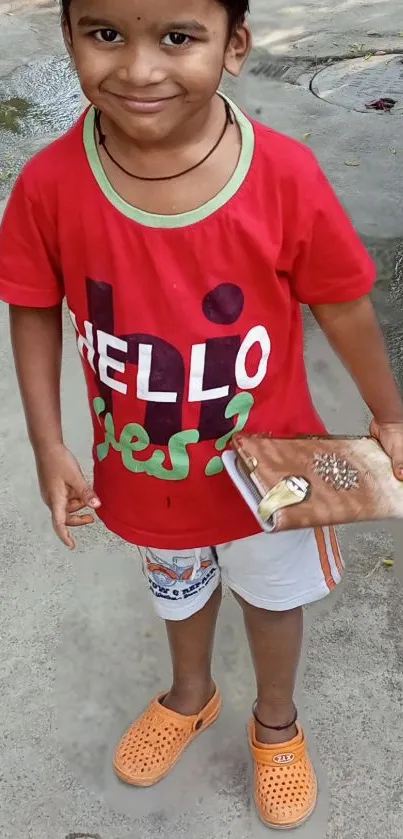 A cheerful child in a vibrant red shirt holding a clutch, standing on pavement.