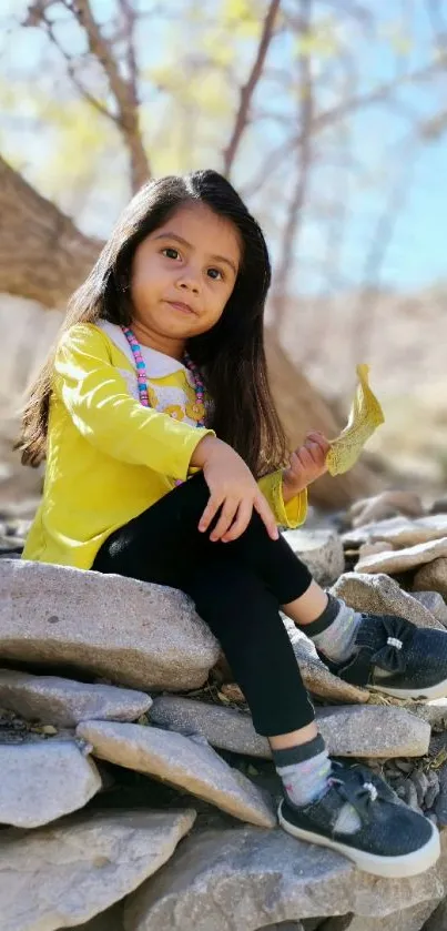 Young girl sitting on stones in sunlight wearing a yellow shirt.