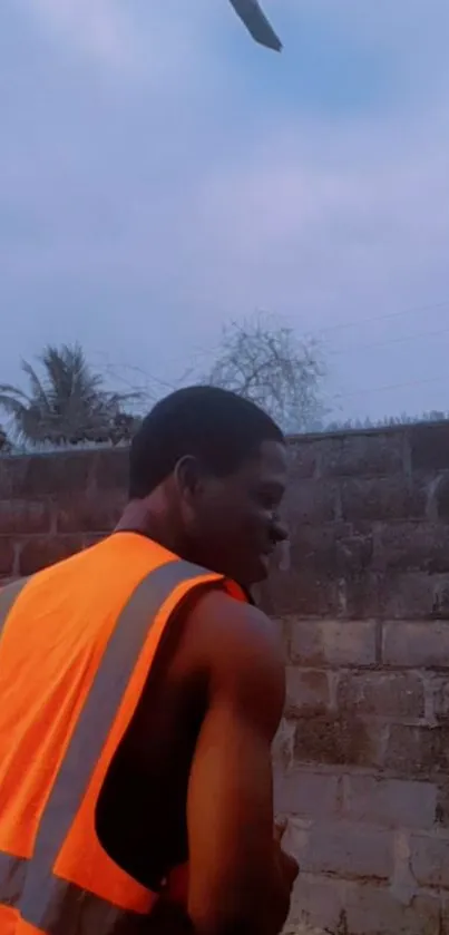 Person wearing an orange vest against a brick wall at twilight.