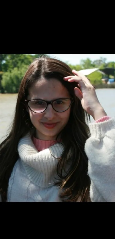 Woman with glasses smiling by a river in a cozy sweater.