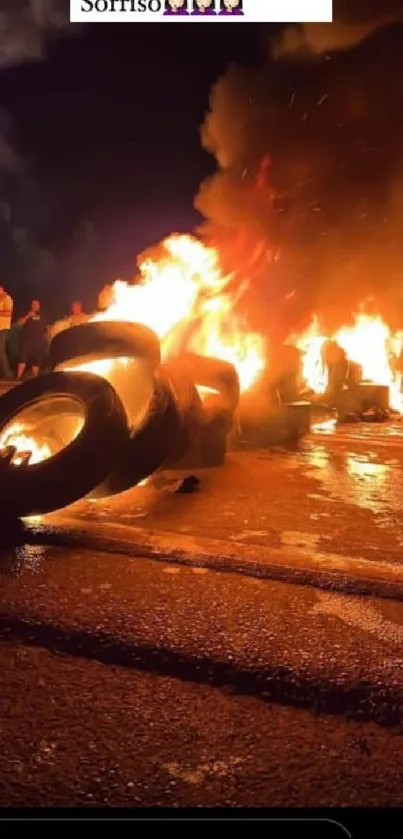 Burning tires on a dark road at night, with vivid flames and onlookers present.