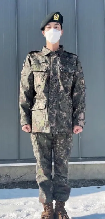 Person in camouflage uniform stands on snow with a gray industrial background.