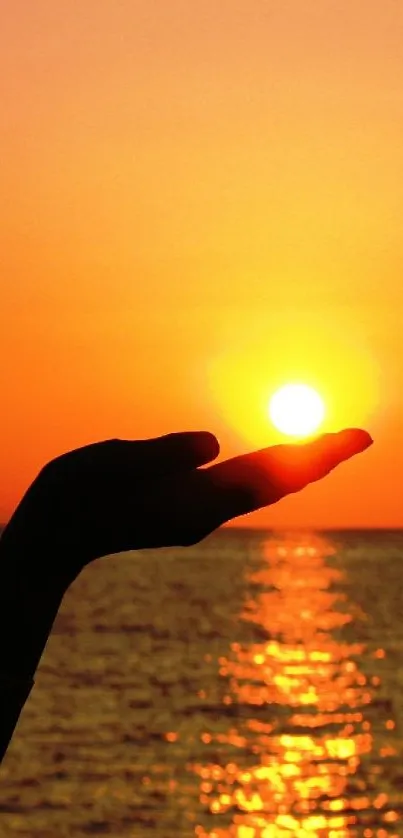 Silhouette of a hand holding the sun against an orange sunset over the ocean.