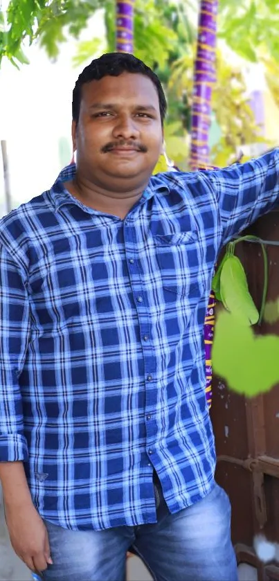 Man in blue plaid shirt stands outdoors, surrounded by greenery.