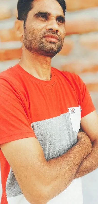 Man in red shirt against brick wall, arms crossed.