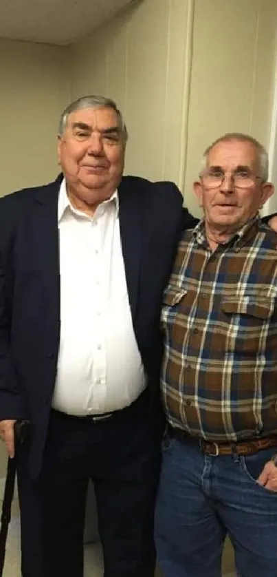 Two elderly friends posing for a casual portrait against a beige wall.