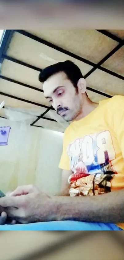 Man in a yellow shirt relaxing with phone under a bed.