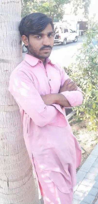 Man in pink traditional outfit leans against a tree in daylight.