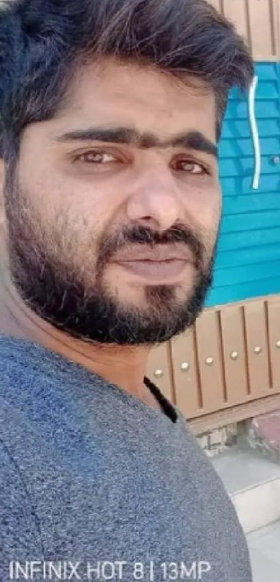 A man with a beard poses outdoors in front of a wooden and turquoise shutter.