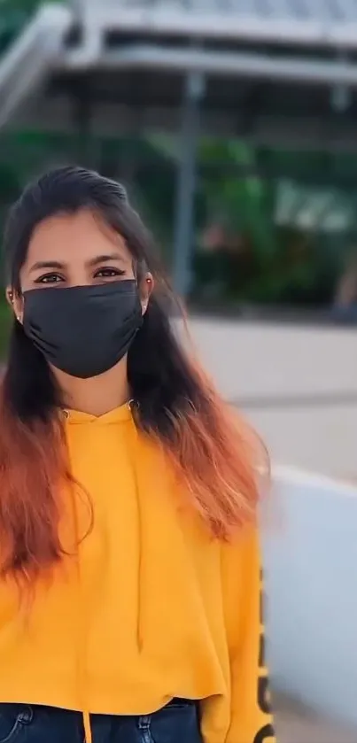 A young woman wearing a yellow hoodie and mask outdoors.