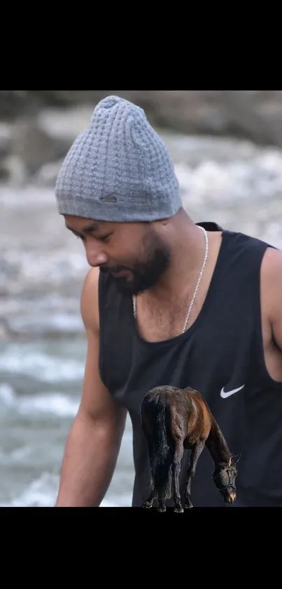 A person in a black tank top and gray beanie beside a river, with a horse.