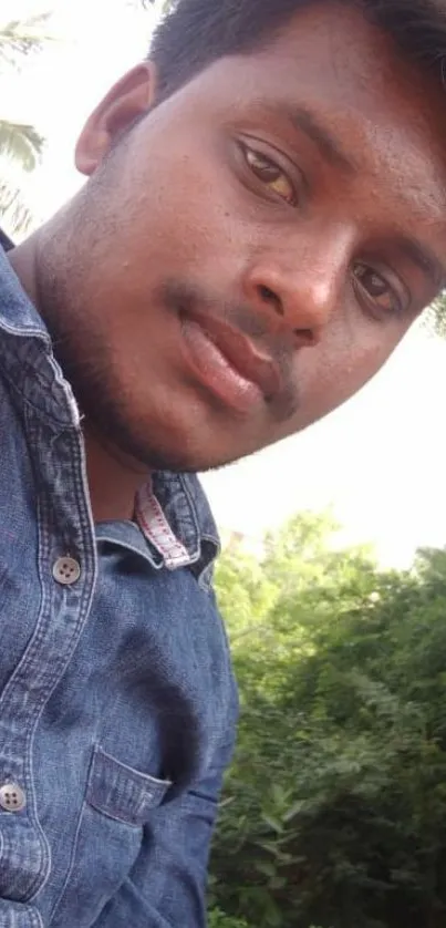 Young man in denim shirt outdoors with greenery.