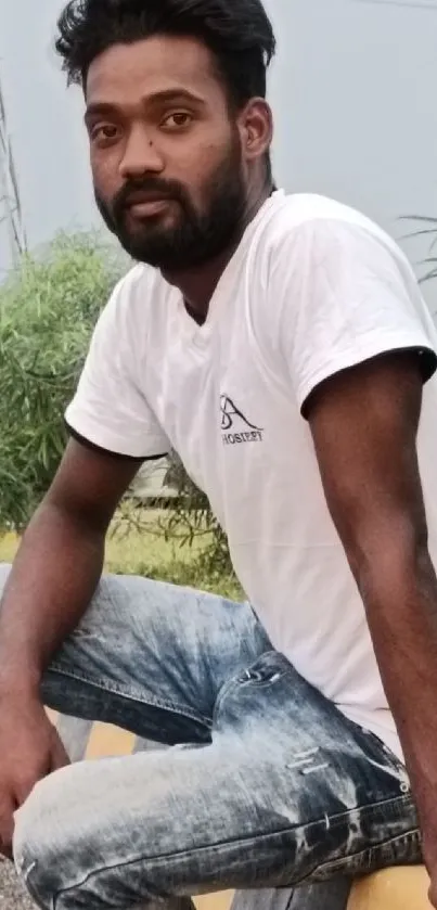 Young man in casual attire sitting outdoors with greenery.