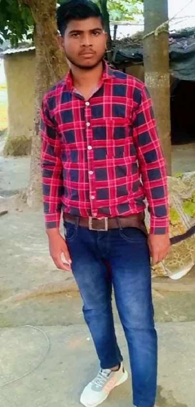 Young man in red plaid shirt outdoors in a rural setting, standing on a sunny day.