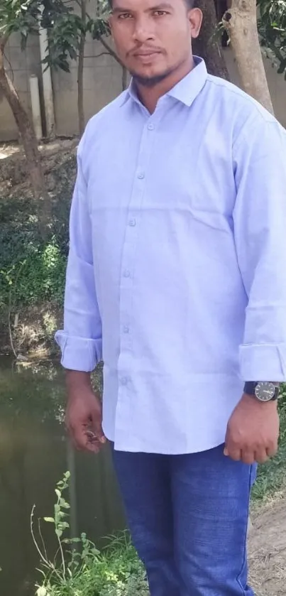 Man in blue shirt standing by trees and pond.