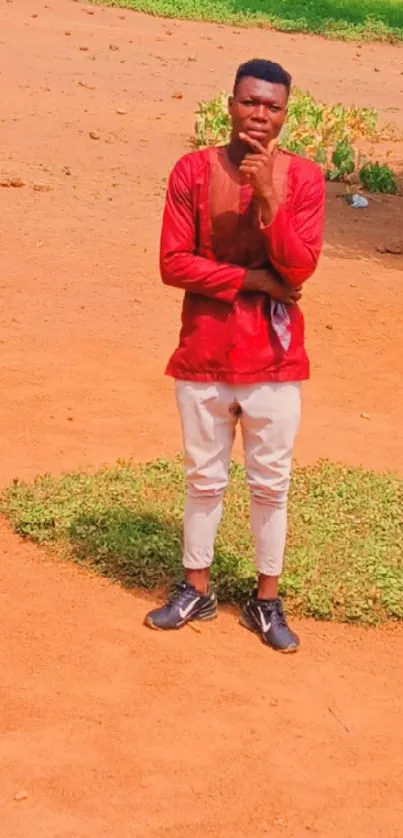 Man in red shirt standing outdoors on sandy ground.