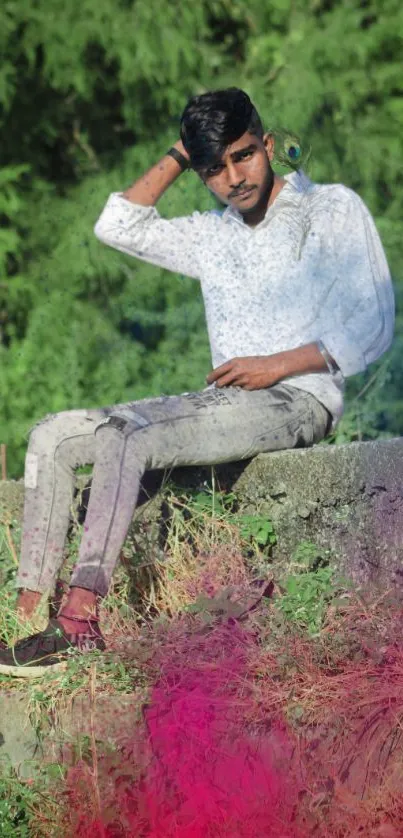 Young man in casual attire sitting on a wall, surrounded by green leaves and pink hues.