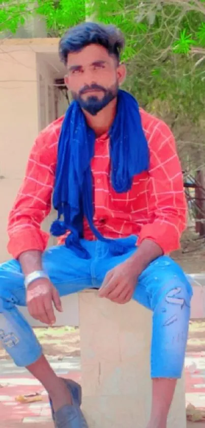 Man in red shirt and blue jeans sitting outdoors.