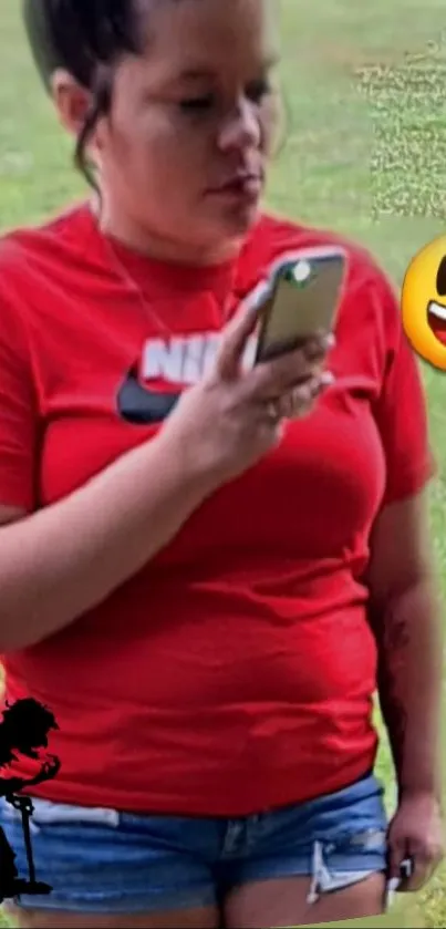 Woman in red shirt checking phone outdoors with emoji.