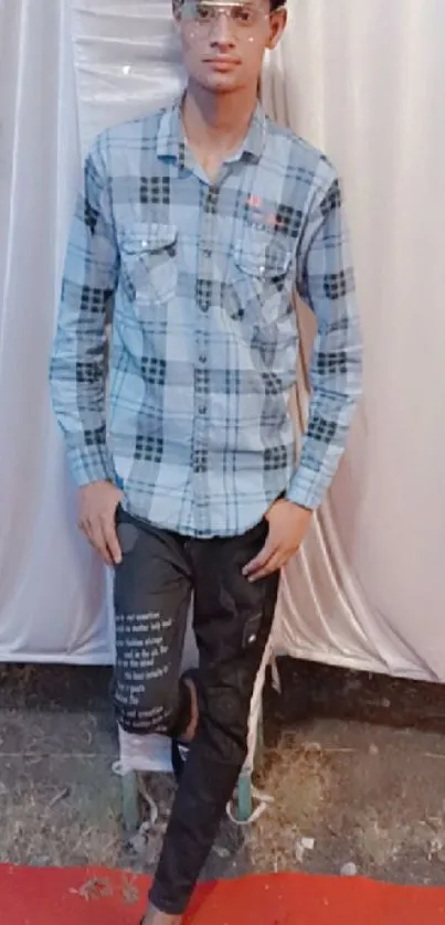 Young man in blue plaid shirt and black jeans standing against a white background.