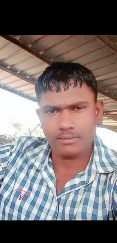 Man in blue checkered shirt sitting casually under a roof.