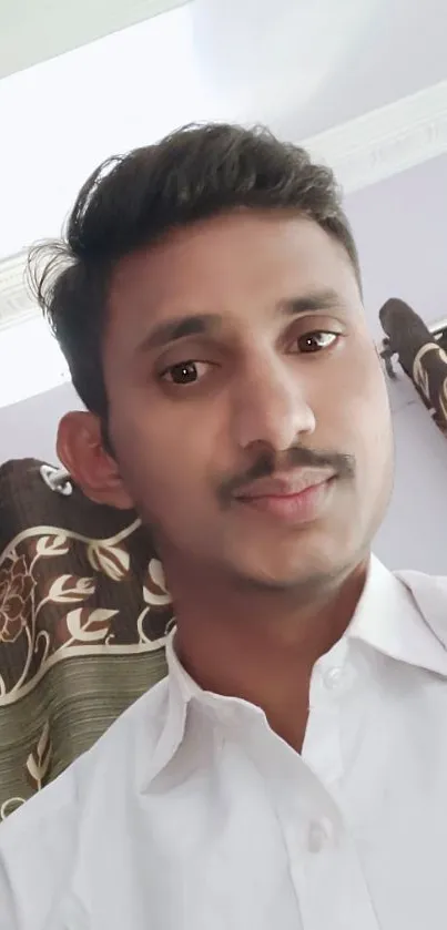 Portrait of a young man in a white shirt with floral curtains in the background.